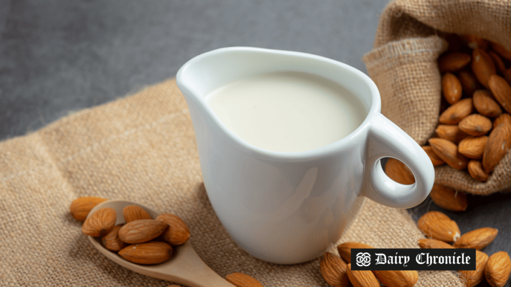 A cup of almond milk on a table, surrounded by scattered almonds and a spoon filled with almonds.