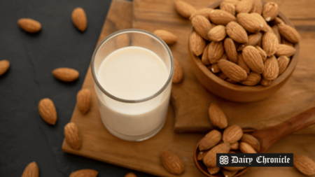 A glass of almond milk beside a bowl full of almonds, with some almonds spread on the table